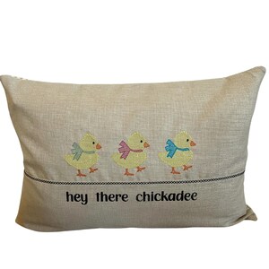 May include: A beige pillow with embroidered yellow chicks wearing bows. The text "hey there chickadee" is embroidered below the chicks.