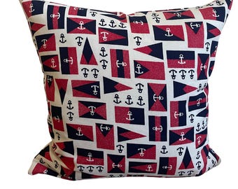 May include: Square decorative pillow with a nautical theme. The pillow features a repeating pattern of red, navy blue, and white nautical flags and anchors. The pillow is square and appears to be made of a soft fabric.