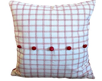 May include: Square pillow with a white background and a red grid pattern. The pillow has five red buttons along the bottom edge. The pillow is a decorative home accent.