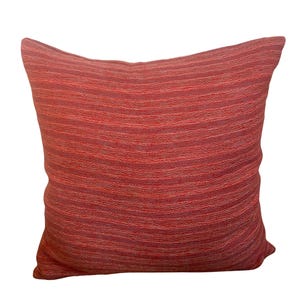 May include: A square decorative pillow in shades of red. The pillow has a textured, woven appearance with horizontal stripes. The pillow is a solid color and appears to be made of a soft fabric. The pillow is a home decor item.