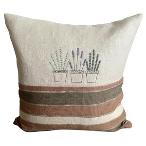 May include: Cream-colored decorative pillow with embroidered herbs. The top section features rosemary, lavender, and thyme in pots. The bottom has horizontal stripes in brown, tan, and cream. A cozy accent for a living room or bedroom.