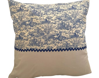 May include: A decorative throw pillow with a blue and white toile pattern featuring a cottage scene. The pillow has a beige backing and a blue scalloped trim.