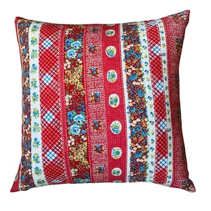 May include: Square decorative pillow with a patchwork design. The pillow features vertical stripes of red, white, and blue plaid, floral patterns, and solid red sections. The floral patterns include blue, yellow, and white flowers.