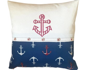 May include: A white pillow with a red and white striped anchor embroidered on the front. The back of the pillow is blue with a pattern of white and red anchors.