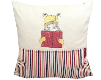 May include: A cream-colored pillow with a pocket on the bottom. The pocket is striped in red, yellow, blue, and white. The pillow features an embroidered design of a girl wearing a yellow hat reading a red book.