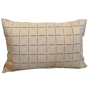May include: Rectangular decorative pillow with a cream-colored background and a grid pattern of thin, dark red lines. The pillow has a soft, textured appearance, suggesting a natural fabric. The pillow is designed for home decor.