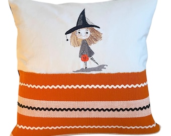 May include: White pillow with a black and orange striped border. The pillow features an embroidered image of a girl dressed as a witch holding a pumpkin bucket.
