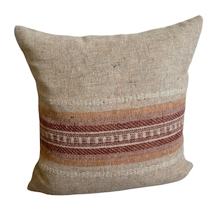 May include: Square beige pillow with a woven design. The pillow features horizontal bands of orange, brown, and white. The pillow is made of a textured fabric, suitable for home decor.