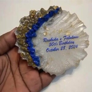 May include: A decorative, irregularly shaped resin piece with a white and gold base, a blue river-like accent, and the inscription "Rashida's Fabulous 50th Birthday October 28, 2024" in blue script. Gold flecks add detail.