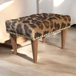 May include: A leopard print upholstered ottoman with four wooden legs and gold nailhead trim.