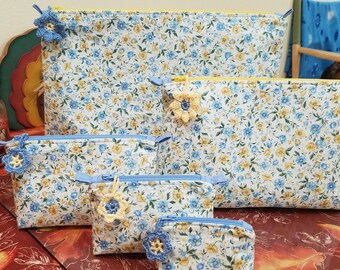 May include: Five zippered pouches in various sizes, all made of white fabric with a blue and yellow floral pattern. The pouches have blue zipper pulls and blue crocheted flower embellishments.