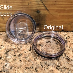 May include: Two clear plastic lids for a travel mug. One lid has a slide lock feature, the other is a standard lid. The lids are on a granite countertop.