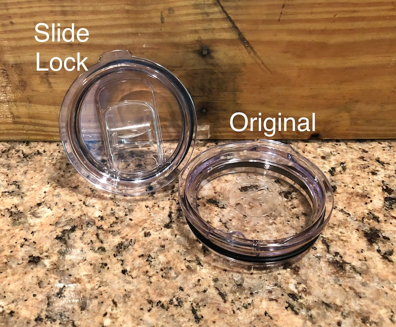 May include: Two clear plastic lids for a travel mug. One lid has a slide lock feature, the other is a standard lid. The lids are on a granite countertop.