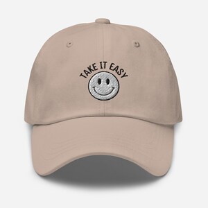 May include: A beige baseball cap with black embroidery that reads "Take It Easy" and a white smiley face.