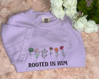 Puede incluir: Una camiseta lavanda con una fila de flores coloridas y el texto "Rooted in Him".