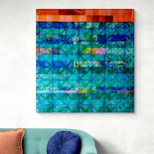 May include: A square wall art piece featuring a grid of blue and teal squares with tie-dye patterns, topped by a horizontal band of orange and red squares. Below the art sits a light teal sofa with a peach square pillow and a round navy pillow.
