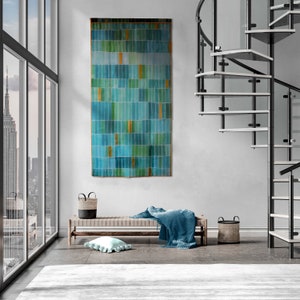 Puede incluir: Un gran tapiz rectangular que presenta una cuadrícula de segmentos verticales de color azul verdoso, azul y verde con acentos amarillos ocasionales. La pieza textil tiene un borde oscuro y cuelga plana contra una pared gris claro.