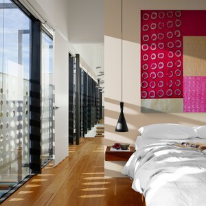 May include: A modern bedroom with a white bed, white bedding, and a large abstract painting with pink and red circles. The room has a large window with a view of a building with a patterned facade.