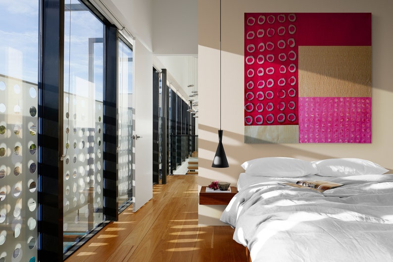 May include: A modern bedroom with a white bed, white bedding, and a large abstract painting with pink and red circles. The room has a large window with a view of a building with a patterned facade.