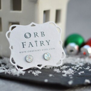Peut inclure: Une paire de boucles d'oreilles clous argentées avec des paillettes irisées sur une carte blanche avec le texte "ORB FAIRY" et l'adresse du site Web "www.orbfairy.etsy.com".
