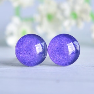 Peut inclure: Deux boucles d'oreilles rondes de couleur violette avec un effet scintillant. Elles sont présentées sur un fond flou avec des fleurs blanches et le texte "ORBFAIRY.ETSY.COM" en haut.