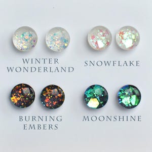 May include: A display of six pairs of round stud earrings from "The Winter Collection." The earrings are on a white card with text labels: "Winter Wonderland," "Snowflake," "Burning Embers," and "Moonshine." The website address is visible at the bottom.