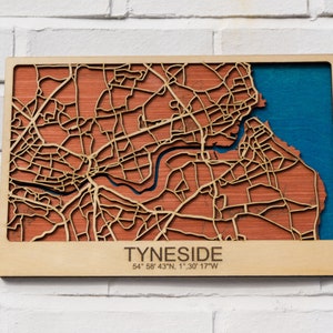 May include: A wooden map of the city of Tyneside, with a blue water feature and the coordinates 54° 58' 43" N, 1° 30' 17" W. The map is mounted on a light wood frame.