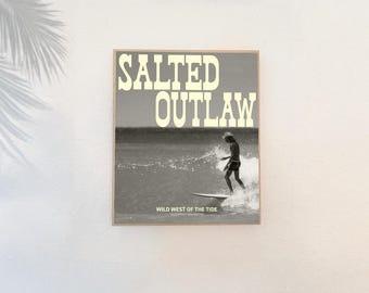 May include: A framed sepia-toned print featuring a surfer riding a wave. The print has the words "SALTED OUTLAW" in large, bold letters at the top, with "WILD WEST OF THE TIDE" at the bottom. The image is set against a white wall.