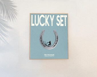 May include: A framed art print with the words "LUCKY SET" in white block letters. A silver horseshoe frames a silhouette of a surfer on a light blue background. The text "TIDE WRANGLER" and "MASSIMO COLLECTIVE" are below the horseshoe.