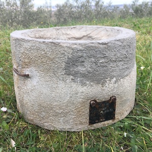 May include: A round, grey concrete planter with a metal ring handle and a metal plaque attached to the bottom. The plaque has the text "2708 1960 1970" on it.