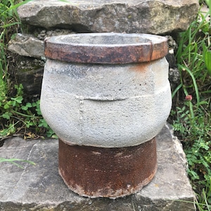 Może przedstawiać: Zwietrzała, szara betonowa donica z rdzawym metalowym pasem wokół górnej krawędzi. Donica ma pękaty korpus i stoi na cylindrycznej, rdzawej podstawie. Tekstura jest szorstka i postarzała.