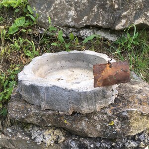 Może przedstawiać: Betonowa kąpiel dla ptaków z przymocowaną z boku rdzewiejącą metalową etykietą. Kąpiel dla ptaków stoi na stercie kamieni.