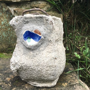 Könnte beinhalten: Ein handgefertigter, grauer Beton-Eulen-Blumentopf mit einem herzförmigen Mosaikauge. Das Mosaik zeigt blaue, braune und grüne Fliesen. Er hat einen Metallgriff und steht auf einer Steinoberfläche, umgeben von Grün.