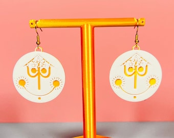 May include: A pair of white earrings with a gold design of a tree with three suns on a stand. The stand is orange and has a hexagonal base.