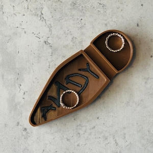 Puede incluir: Un joyero de madera con la palabra "ANDY" tallada en él. Dos anillos de oro se colocan en el joyero.