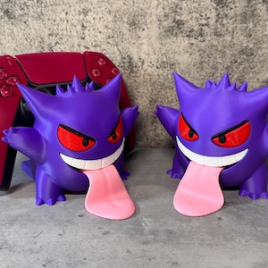 May include: Two purple and black 3D printed Ghost Pokemon figures with red eyes and pink tongues. The figures are holding a red video game controller.