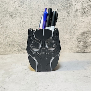 May include: Black plastic desk organizer shaped like the Black Panther mask. The organizer has white details and is holding three pens.