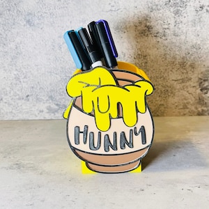 May include: Yellow honey pot shaped desk organizer with a dripping honey design and the word "Hunny" on the front. The organizer has four black and blue pens inside.