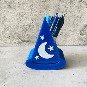 May include: A blue 3D printed pen holder shaped like a wizard's hat with a crescent moon and stars design. The holder is filled with pens and pencils.