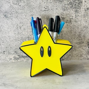 Peut inclure: Un organisateur de bureau en forme d'étoile jaune avec une bordure noire et un motif d'œil noir et blanc. L'organisateur est rempli de stylos et de marqueurs.