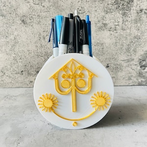 May include: White ceramic pencil holder with a gold design of a stylized tree with two suns on either side. The holder has a small hole in the bottom for drainage.