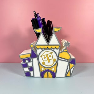May include: A whimsical pen holder shaped like a colorful castle, featuring white, yellow, purple, and gray accents. The castle design includes a smiling face and holds several pens and markers. The background is a solid pink.