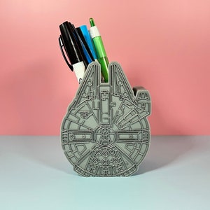 Op de afbeelding: Een grijze Millennium Falcon-vormige bureau-organizer met een zwarte, blauwe en groene pen erin.