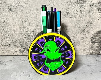 Puede incluir: Un organizador de escritorio negro y verde con forma de rueda con un borde morado y amarillo. El centro de la rueda presenta una cara verde y negra de Oogie Boogie de la película Pesadilla antes de Navidad.