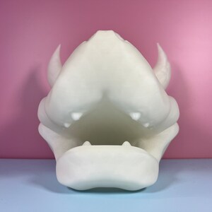Puede incluir: Escultura de resina blanca impresa en 3D de una cabeza de drag&oacute;n con la boca abierta y dientes afilados. La escultura est&aacute; sobre una superficie azul claro.