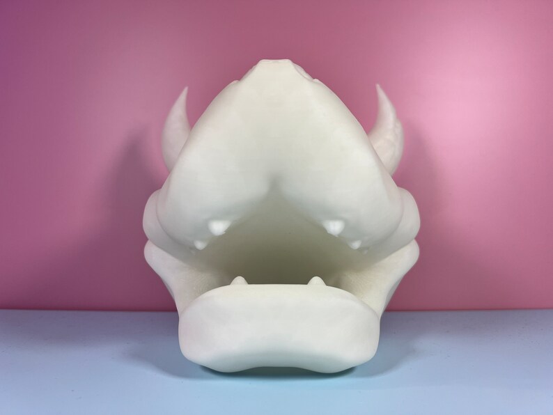 Puede incluir: Escultura de resina blanca impresa en 3D de una cabeza de drag&oacute;n con la boca abierta y dientes afilados. La escultura est&aacute; sobre una superficie azul claro.