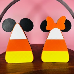 Könnte beinhalten: Zwei Halloween-Dekorationen im Candy-Corn-Design mit Mickey-Maus-Ohren. Eine Dekoration hat eine Schleife oben auf den Ohren.