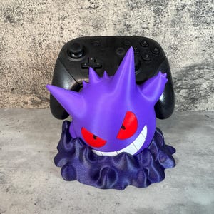 Può includere: Un supporto per controller di videogiochi stampato in 3D di colore viola e nero con un personaggio di Pokémon Gengar.