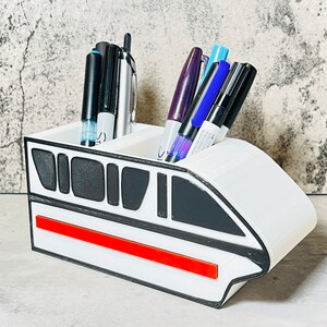 May include: White ceramic pen holder shaped like a futuristic train with a red stripe. The holder has two compartments for pens and pencils.