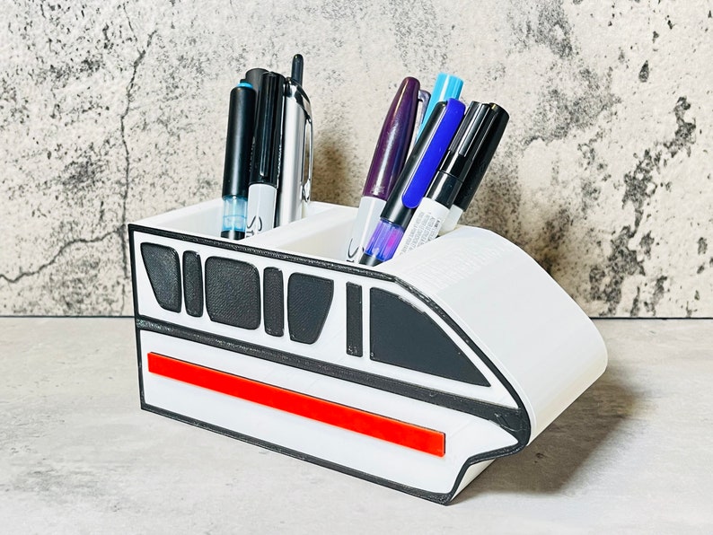 May include: White ceramic pen holder shaped like a futuristic train with a red stripe. The holder has two compartments for pens and pencils.